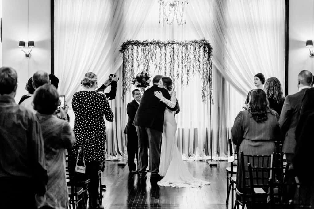 A couple embraces in front of a wedding officiant under a draped floral archway, witnessed by guests seated in rows, in a monochrome setting.