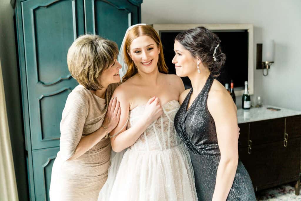 A bride stands between two women, all smiling. The bride wears a white strapless gown. One woman wears a light-colored dress, and the other wears a dark, sparkly dress. They are in a room with a cabinet.