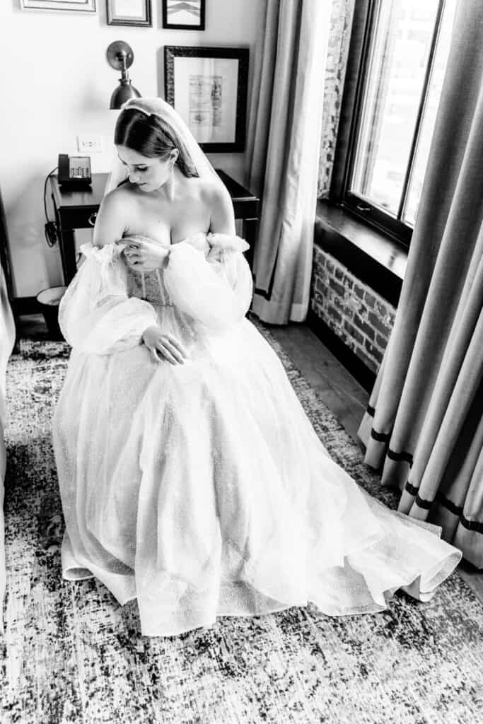 A bride, wearing an off-the-shoulder wedding gown, sits on a chair in a room with a large window, looking down at her dress.