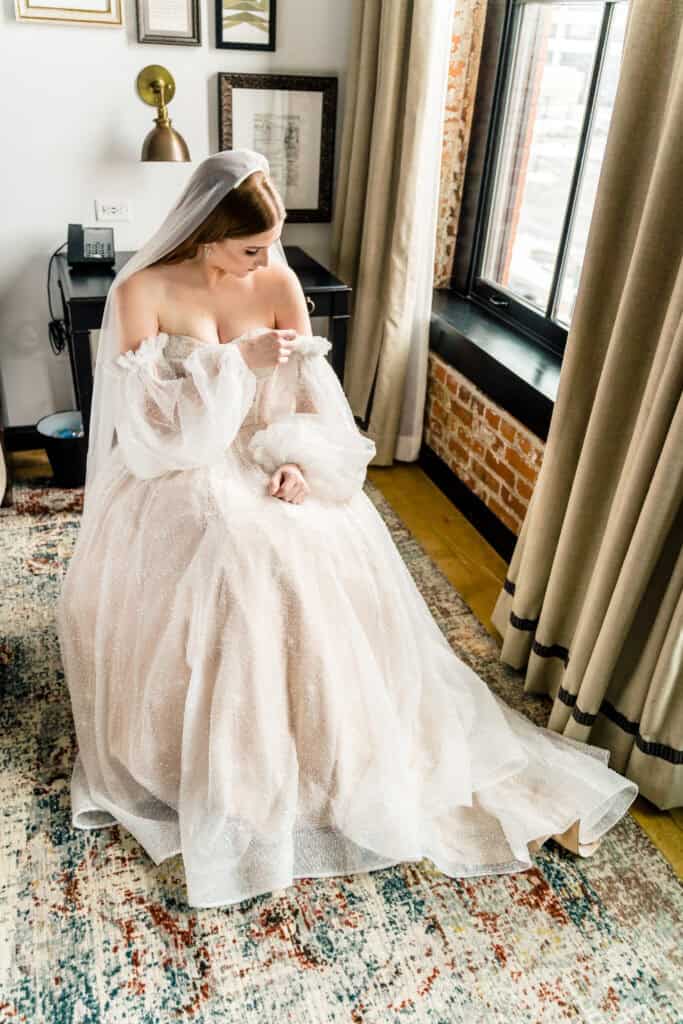 A bride in a white off-shoulder wedding dress with long sleeves sits on a chair, looking down, in a room with a window, curtains, wall decor, and a patterned rug.