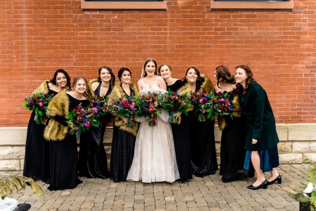 Bride in a white gown with seven bridesmaids in black dresses holding bouquets, posing against a brick wall.