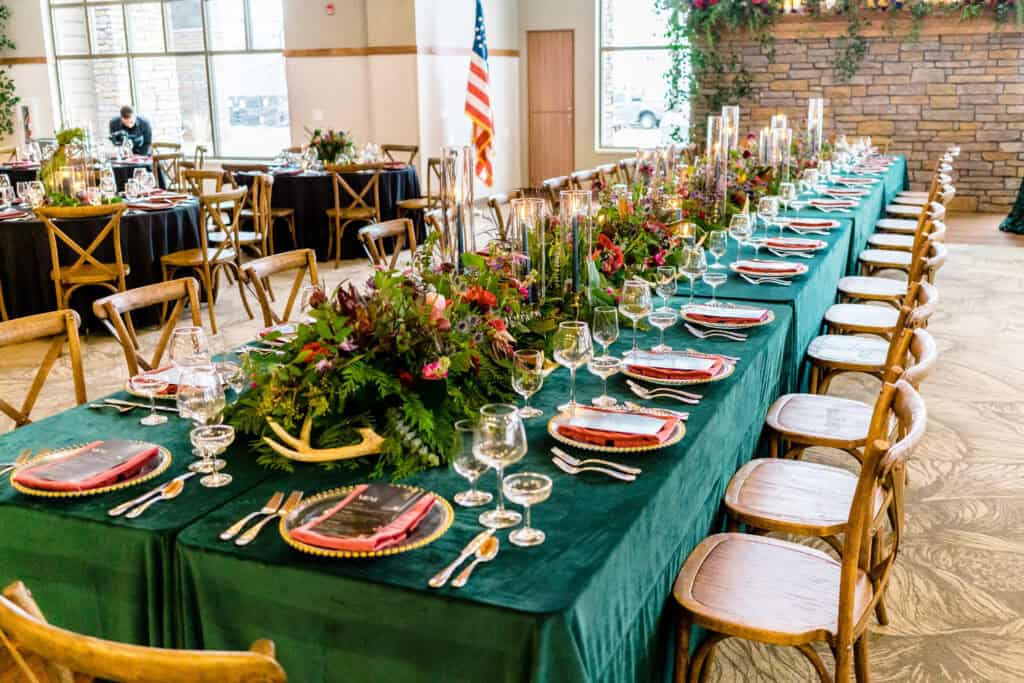 A long dining table set with green tablecloths, red plates, glasses, silverware, and a floral centerpiece in a decorated event space with additional round tables and a US flag in the background.