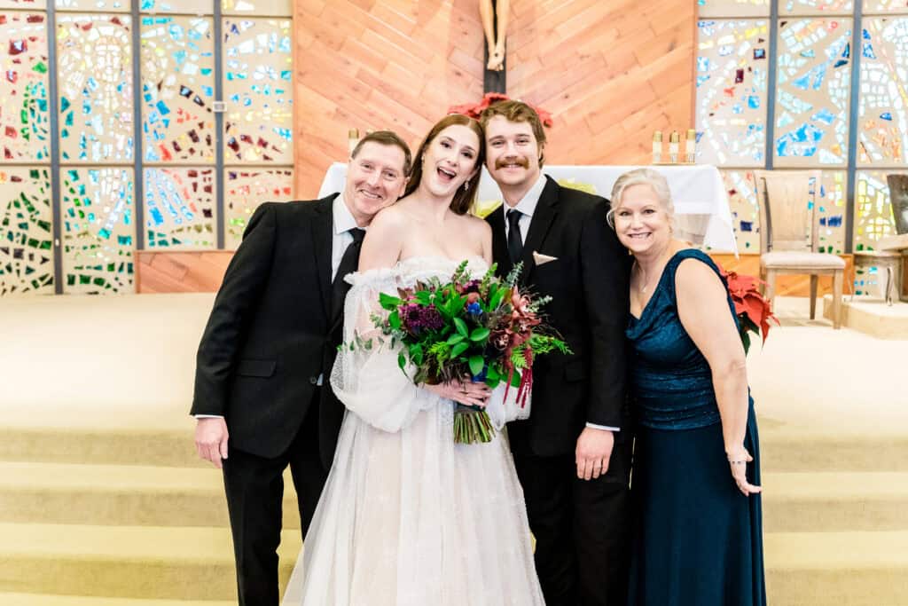 A bride and groom stand with two other adults, all smiling, in front of a colorful stained glass backdrop. The bride holds a flower bouquet.
