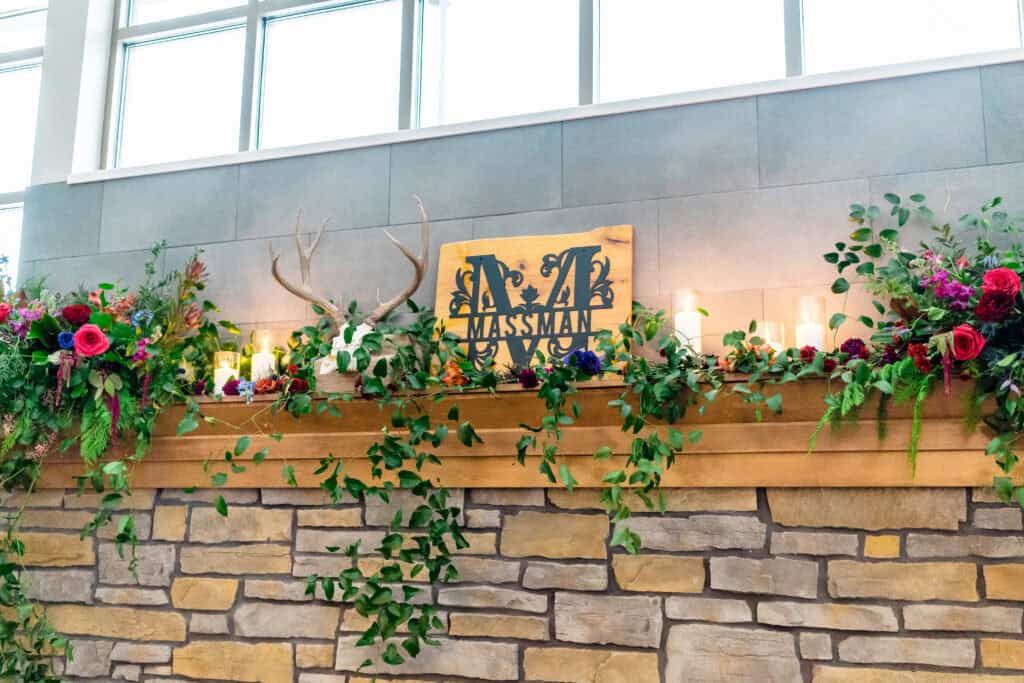Wooden mantelpiece with antlers, a personalized sign, candles, and greenery, adorned with colorful flowers. Brick wall background with large windows above.