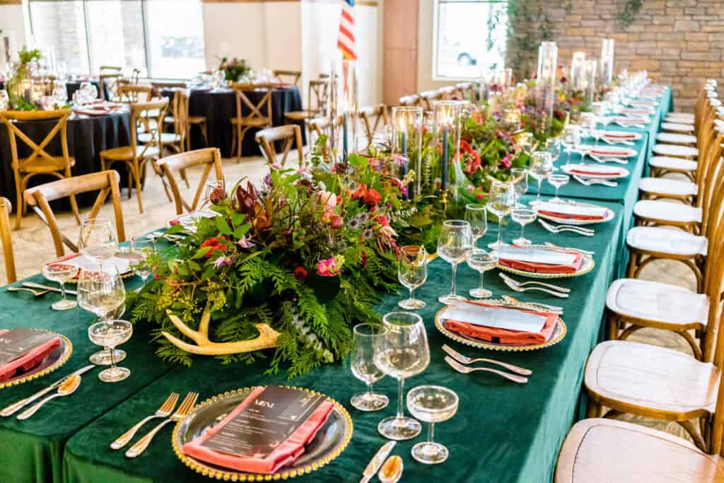 A long dining table adorned with green tablecloths, floral centerpieces, and place settings with glassware and cutlery. Chairs are placed along each side of the table.