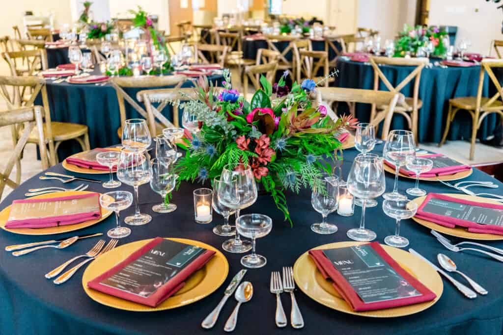 A round table set for a formal event with black tablecloths, gold chargers, red napkins, glassware, silver cutlery, and a centerpiece of vibrant flowers and greenery. Other similar tables are in the background.