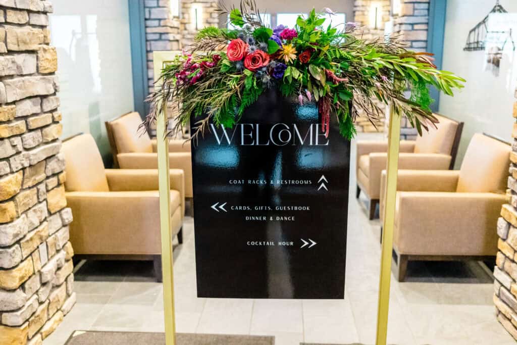 A black welcome sign decorated with flowers, indicating the directions to coat racks, restrooms, cards, gifts, guestbook, dinner and dance, and cocktail hour, in a seating area with beige armchairs.