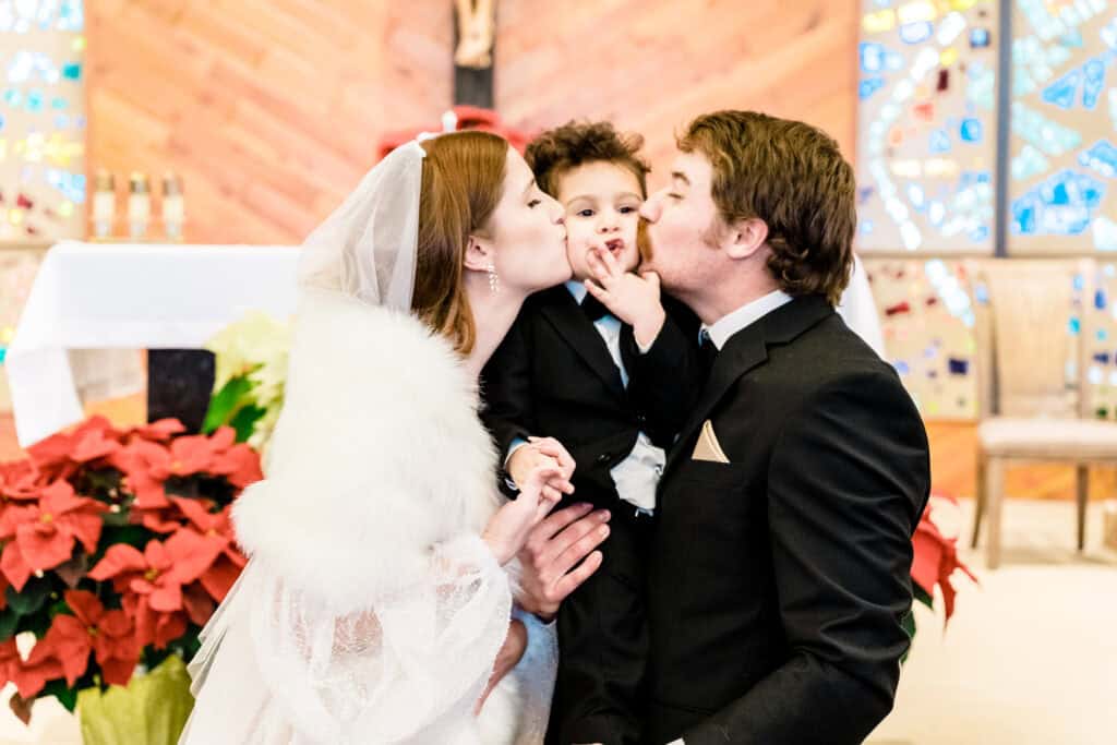 A bride and groom kiss a young child on either cheek while kneeling inside a church. The child, dressed in a suit, touches one adult's nose. The background has a wooden wall and stained-glass windows.