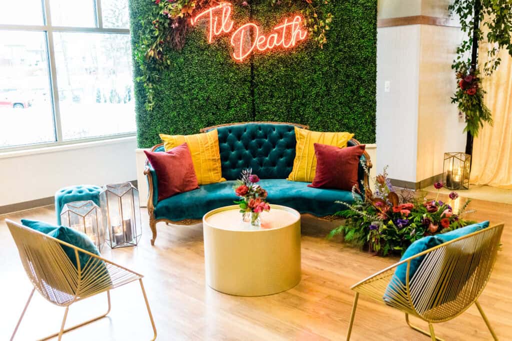 A stylish seating area with a green, cushioned sofa, two wireframe chairs, a round coffee table, and decorative greenery backdrop, featuring a neon sign that reads "Til Death.