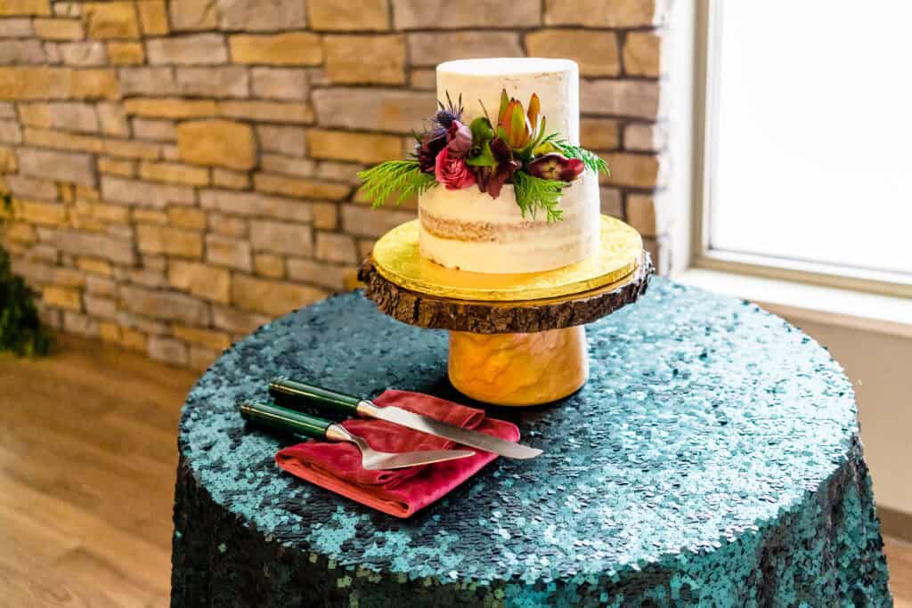 A two-tiered white cake with floral decorations sits on a wooden stand on a sequin-covered table, accompanied by two knives resting on red napkins.