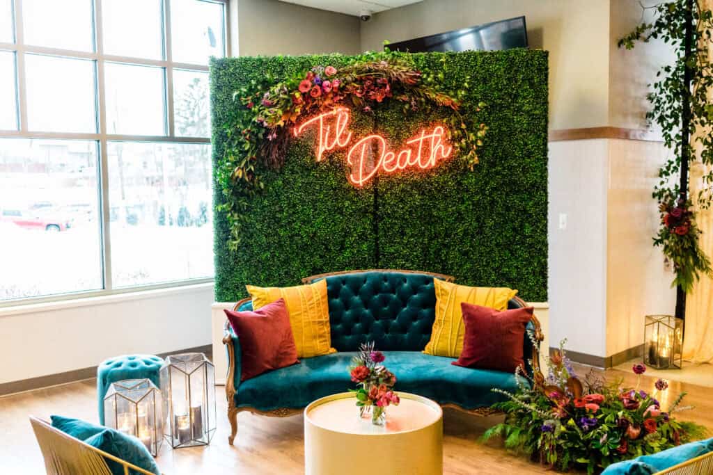A seating area with a green backdrop decorated with flowers and a neon sign that reads "Til Death." The area includes a teal sofa with colorful pillows, a matching ottoman, and decorative lanterns.