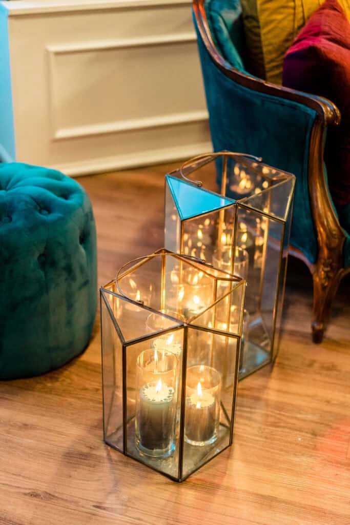 Two glass lanterns with candles on the floor beside a teal armchair and a teal tufted ottoman, creating a cozy ambiance in a room with wooden flooring.