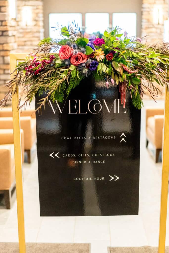 A black welcome sign adorned with a floral arrangement, listing directions to coat racks, restrooms, cards, gifts, guestbook, dinner, dance, and cocktail hour.