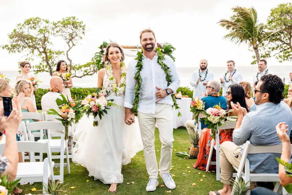 A bride and groom walk down the aisle outdoors, smiling and holding hands. Guests seated on white chairs clap. The setting includes tropical flowers, greenery, and an ocean view in the background.