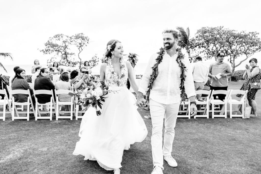 A bride and groom holding hands walk down an outdoor aisle lined with seated guests. The bride is in a white dress holding a bouquet, while the groom wears a lei and white attire.