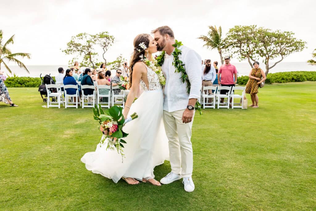 A couple in wedding attire shares a kiss on a grassy area, with guests seated in rows behind them and palm trees in the background. The bride holds a bouquet, and both wear flower garlands.