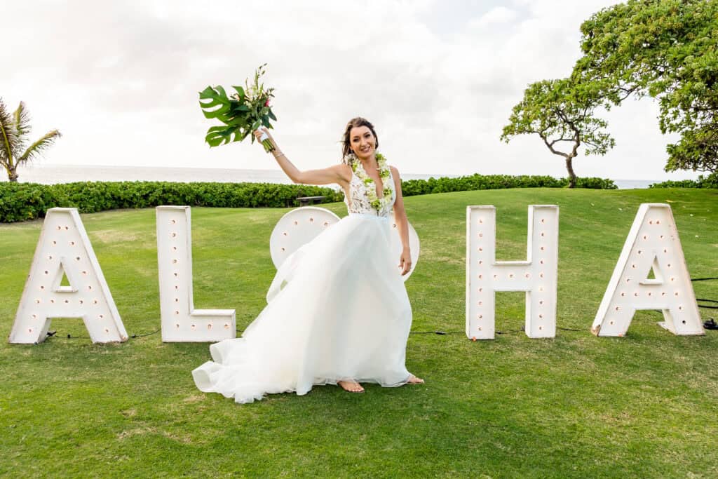 A bride in a white dress holds a bouquet while standing in front of large light-up letters reading "ALOHA," on a grass lawn with trees and ocean in the background.