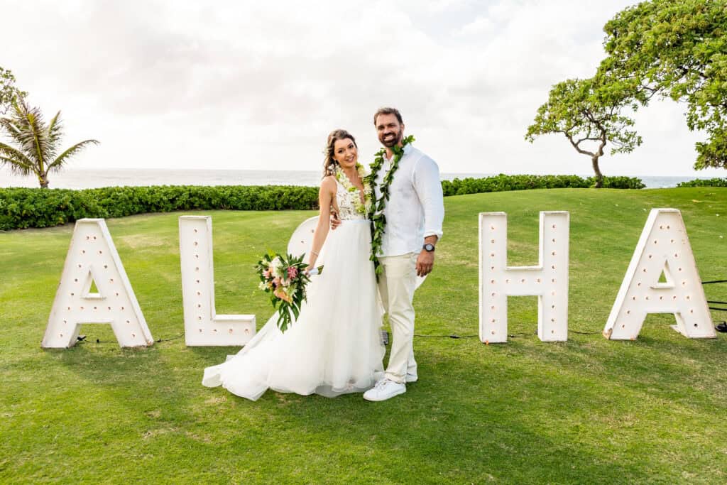 A bride and groom pose together on green grass with large illuminated letters spelling "ALOHA" behind them, trees and the ocean visible in the background.