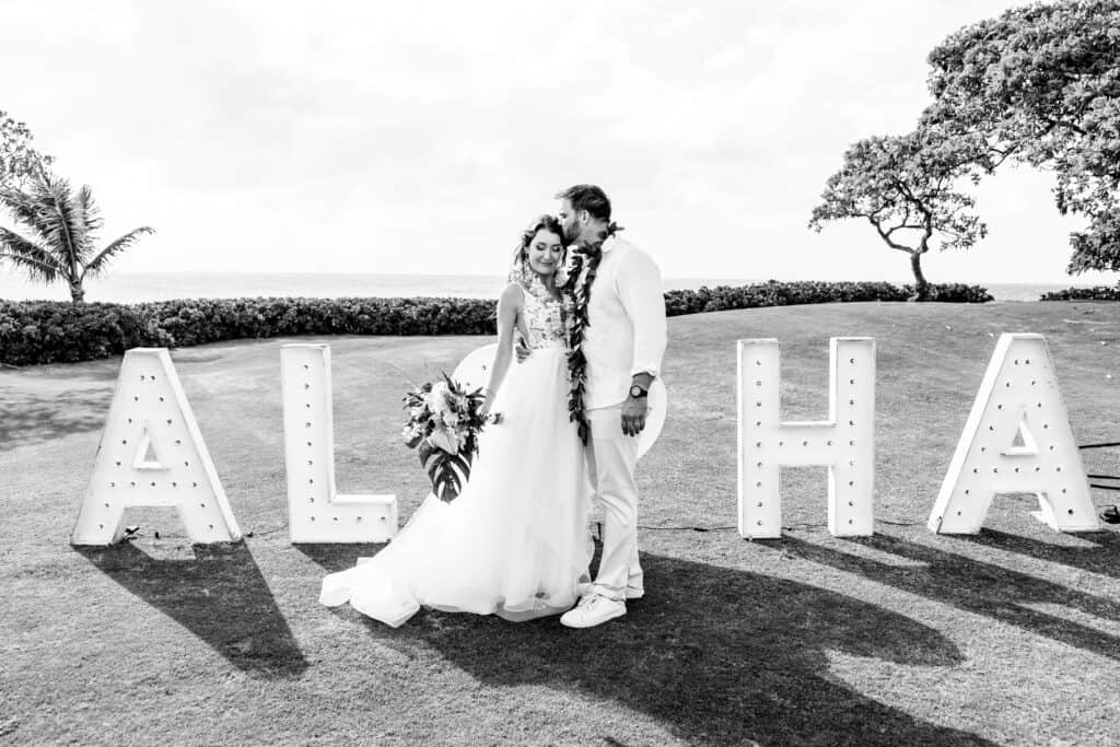 A couple in wedding attire stands in front of large illuminated letters spelling "ALOHA" on a grassy area with trees and ocean in the background.