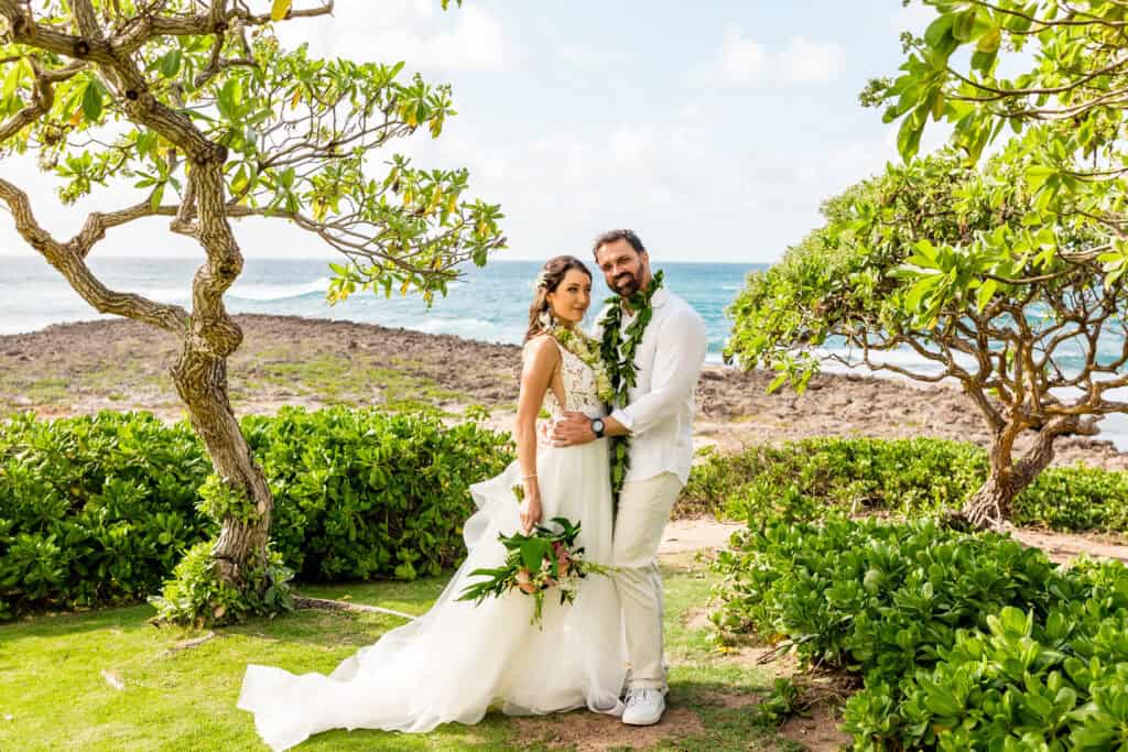 A bride and groom stand together in wedding attire, surrounded by greenery with an ocean backdrop. The bride holds a bouquet, and both are smiling with leis around their necks.