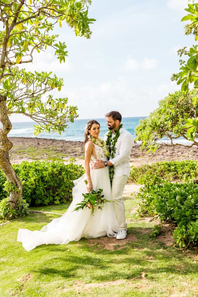 A couple dressed in wedding attire stands together outdoors near the ocean, surrounded by greenery. The bride holds a bouquet and the groom wears a lei.