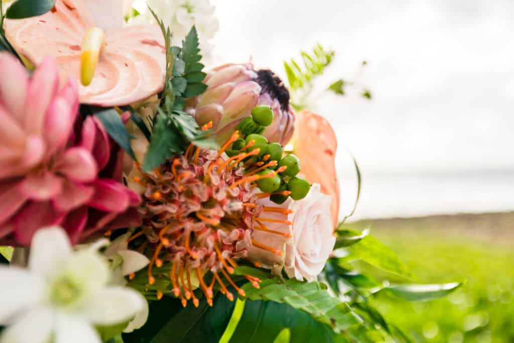 Close-up of a vibrant bouquet featuring various flowers, including white lilies, pink roses, and orange pincushions, with a blurred natural background.