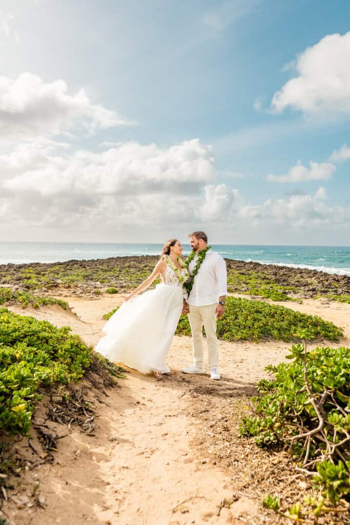 A couple dressed in wedding attire stands on a sandy path surrounded by greenery with the ocean and a partly cloudy sky in the background.