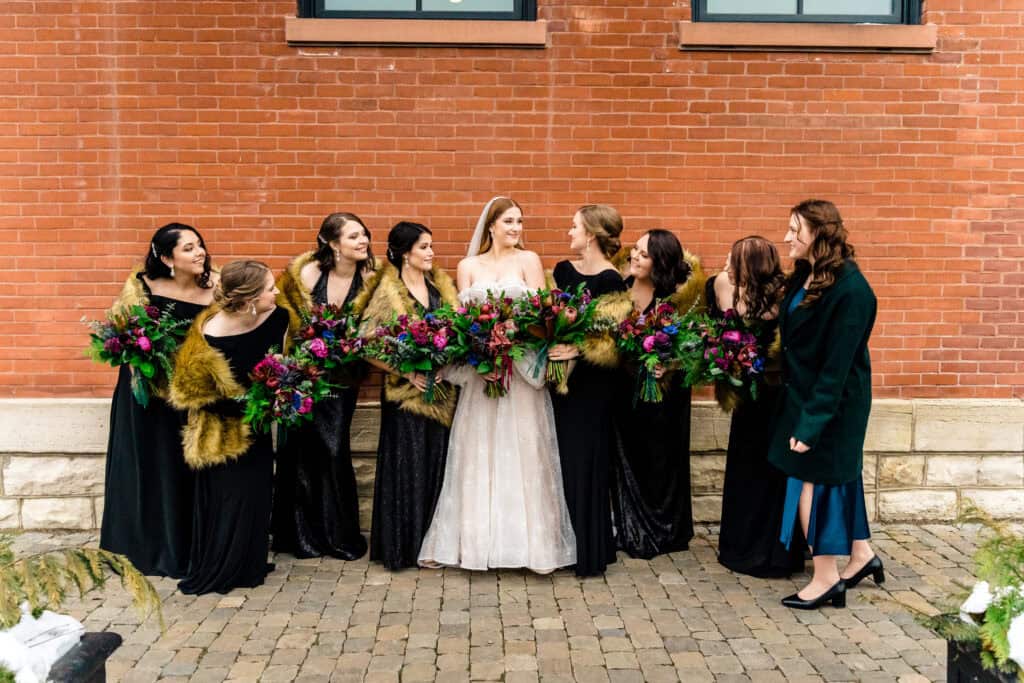 A bride in a white dress stands with six bridesmaids in black dresses holding bouquets, posing in front of a red brick wall. Everyone is looking and smiling at the bride.