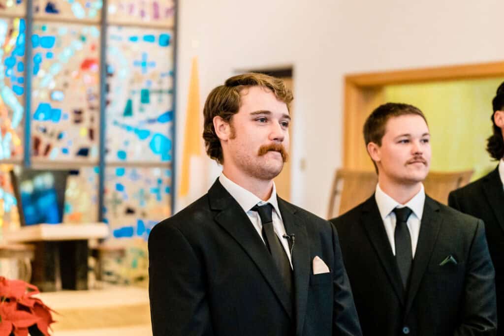 Three men in suits stand indoors, with a stained glass window in the background. Two of the men have mustaches, and they appear to be part of a formal event.