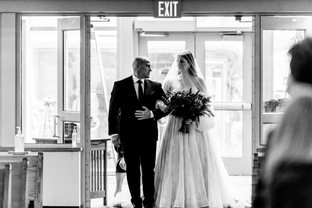 A bride in a white gown holding a bouquet enters a building with a man, possibly her father, who is wearing a suit. They are walking through a doorway under an exit sign.