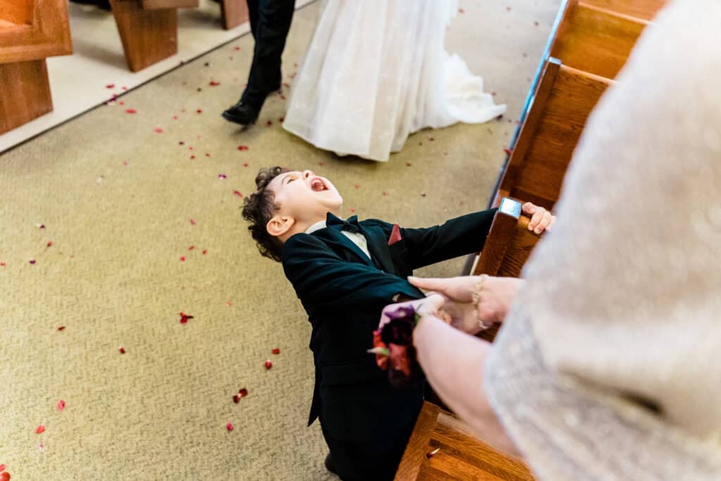 A young boy in a suit is laughing while being guided by an adult in a formal setting with rose petals scattered on the floor.