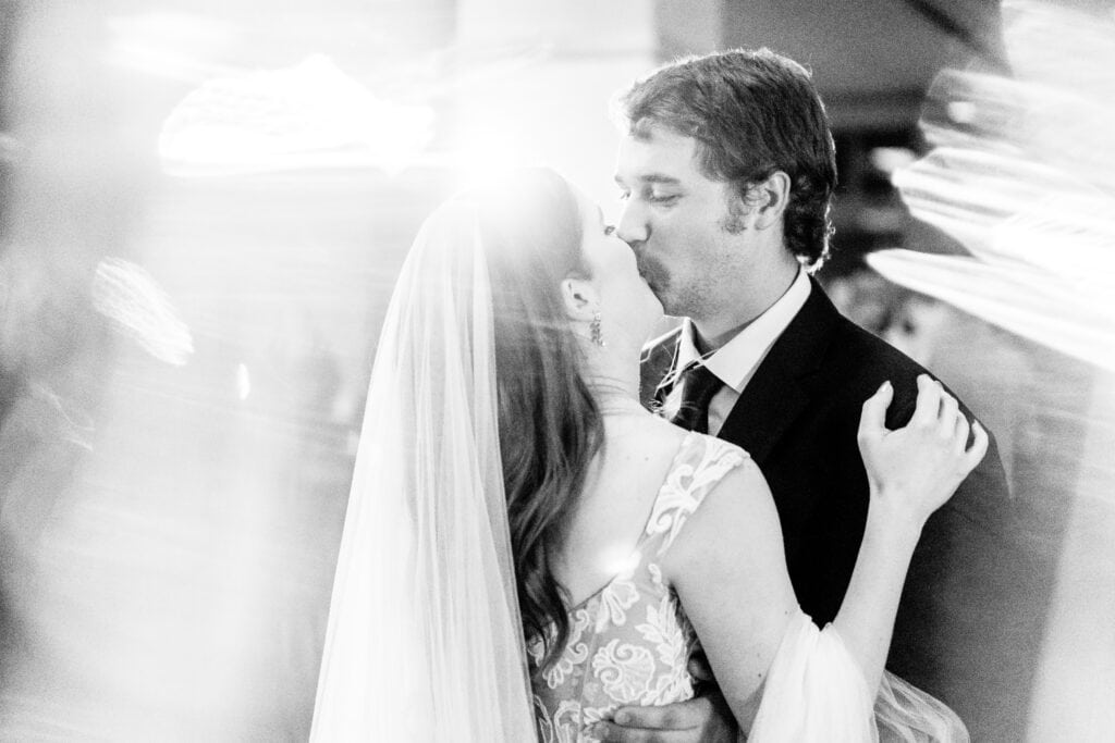 A bride and groom share a kiss on the dance floor, with the bride in a wedding dress and veil, and the groom in a suit. Light streaks create a soft, ethereal effect around them.