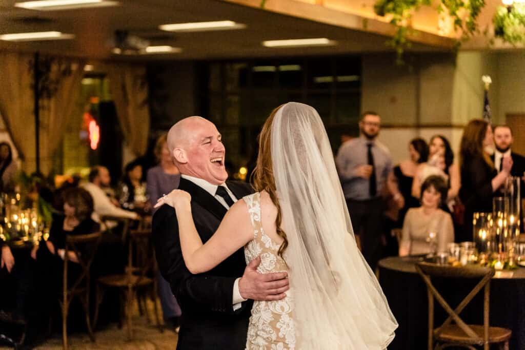 A bride and a bald man in a black suit share a joyful dance at a wedding reception, surrounded by guests seated at tables adorned with candles.