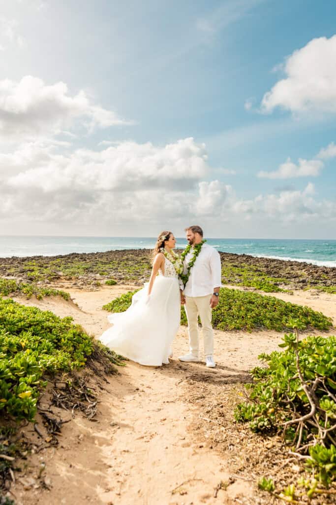 A couple dressed in wedding attire stands on a sandy path surrounded by greenery, with the ocean and a partly cloudy sky in the background.