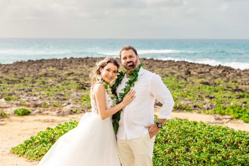 A bride and groom dressed in white, adorned with green leaf garlands, stand together on a rocky coastline with the ocean in the background.