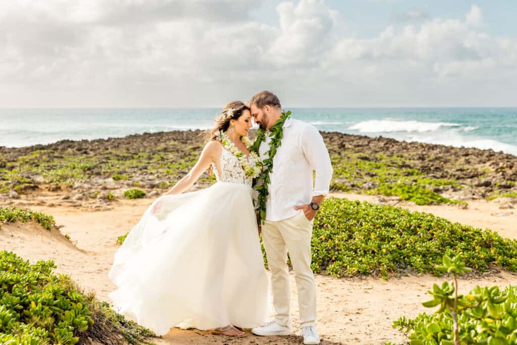 A bride and groom stand close together on a rocky shore with greenery and ocean waves in the background. Both wear white attire, and they are adorned with green leafy garlands.