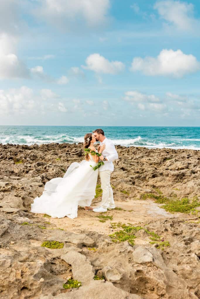 A couple dressed in wedding attire kiss on a rocky shoreline with the ocean and a cloudy sky in the background.
