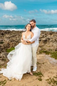 A couple dressed in wedding attire stands embraced on a rocky beach with waves and blue sky in the background.