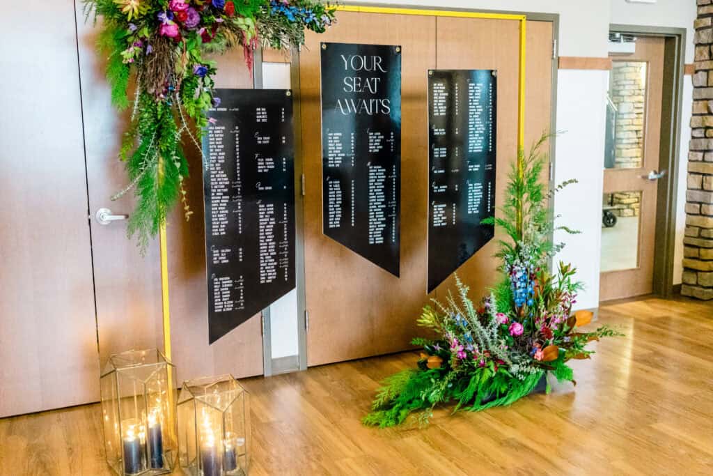 An elegant seating chart display with black panels listing guest names, adorned with floral arrangements and candles, is set against a door with a sign reading, "Your Seat Awaits.