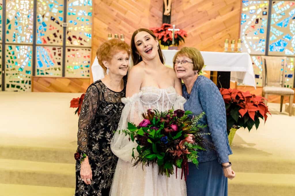 A bride holds a bouquet and poses between two older women in a church setting, all smiling. Stained glass windows are visible in the background.