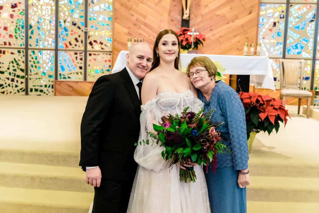 A bride holding a bouquet stands between two smiling people, an older man and an older woman, inside a church with stained glass windows and poinsettias in the background.