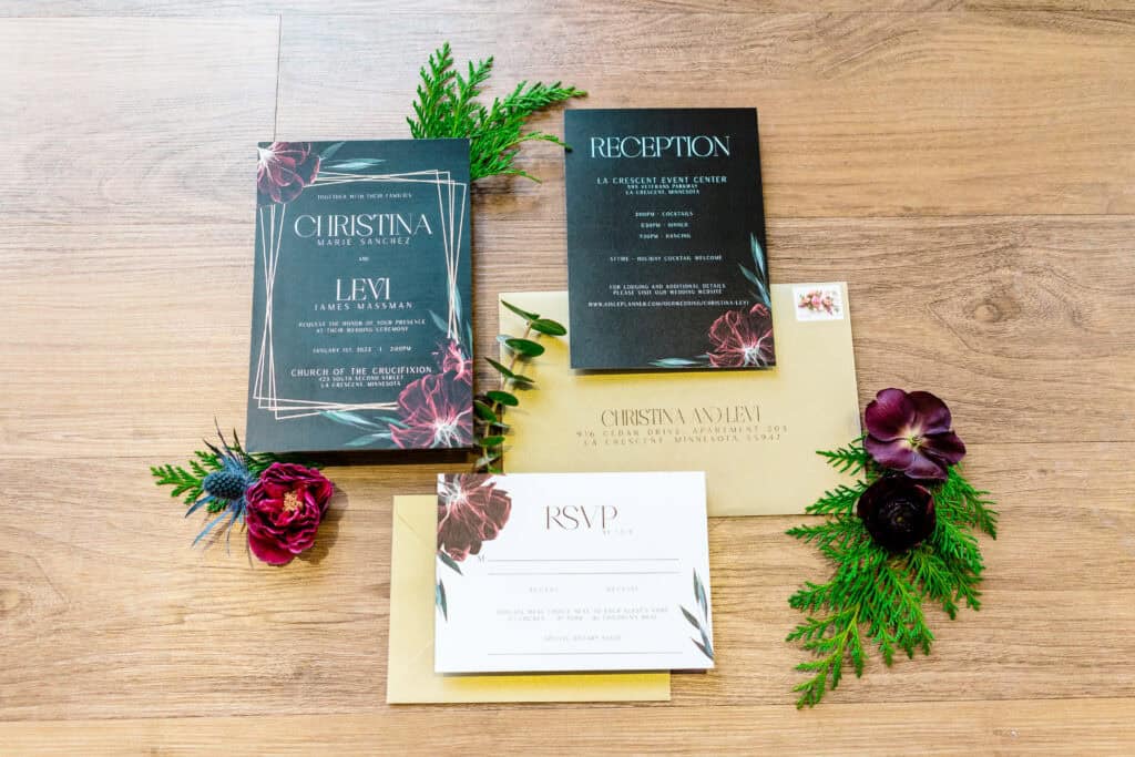 A wedding invitation suite displays black, white, and gold cards labeled "RSVP," "Reception," and "Christina and Levi" with floral accents, set on a wooden surface surrounded by flowers and greenery.