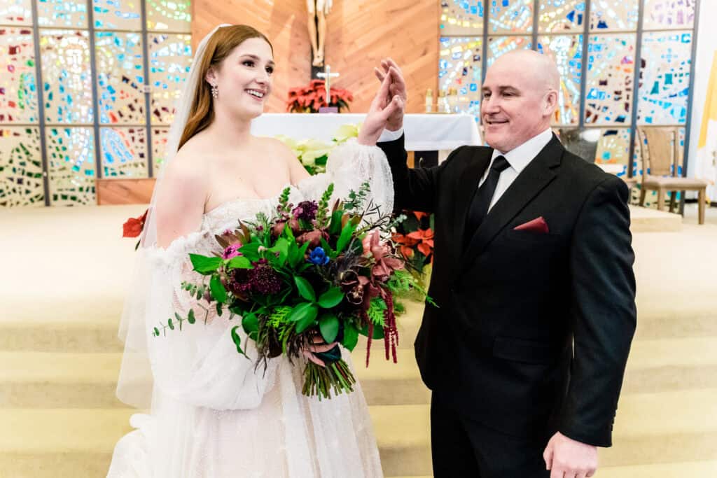 Bride in a white wedding dress holding a bouquet stands next to a man in a black suit inside a decorated venue, both smiling and raising a hand in celebration.