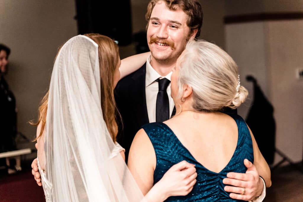 A groom smiles as he embraces a bride and an older woman in a teal dress, possibly during a wedding reception.