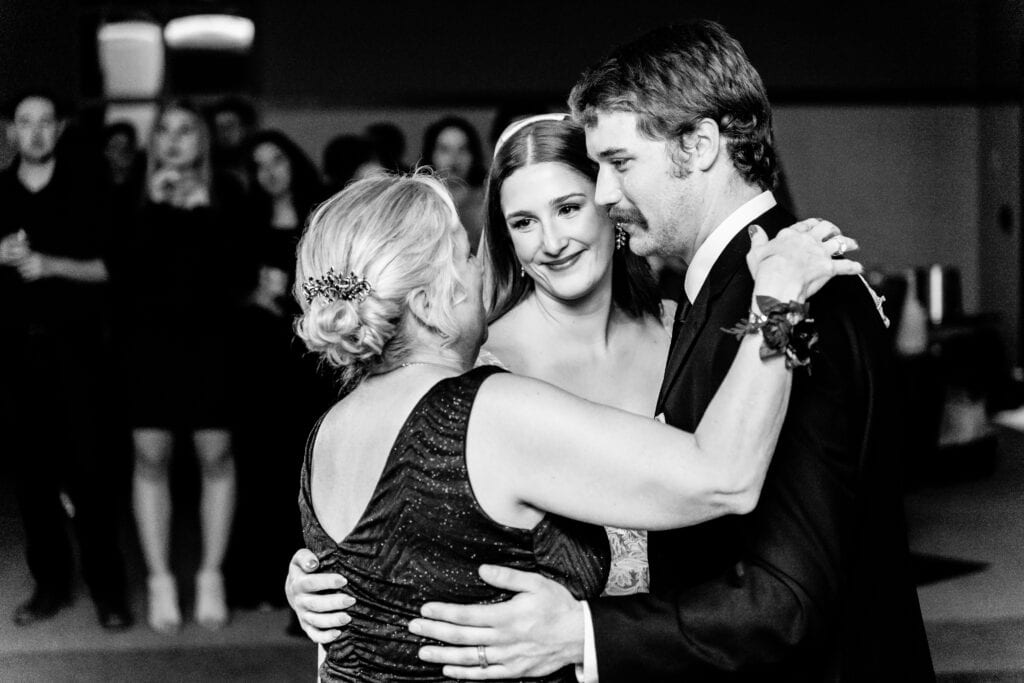 A bride and groom embrace an older woman, all smiling, during an event. Guests in the dimly lit background look on.