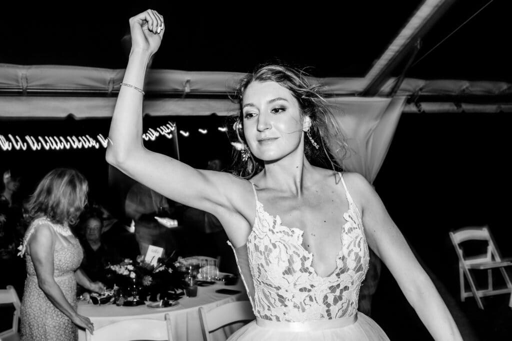 A woman in a sleeveless, lacy dress dances with her arm raised under a tent at a nighttime event. Other people are seen in the background near a table. The image is in black and white.