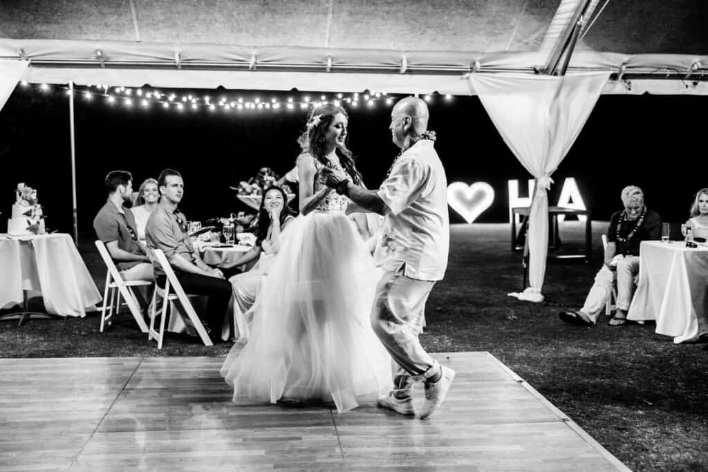 A bride and an older man dance on a wooden floor at an outdoor wedding reception. Guests watch from tables with string lights and illuminated "LA" decor in the background.