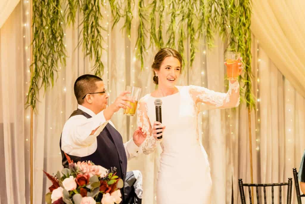 A couple in formal attire raises glasses in a toast, the woman holding a microphone. They stand in front of a backdrop with string lights and greenery. A bouquet of flowers is visible in the foreground.