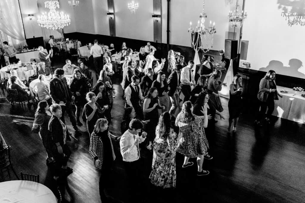 A black and white image of people dancing in a dimly lit ballroom, illuminated by chandeliers. A few tables with seated guests are seen in the background.