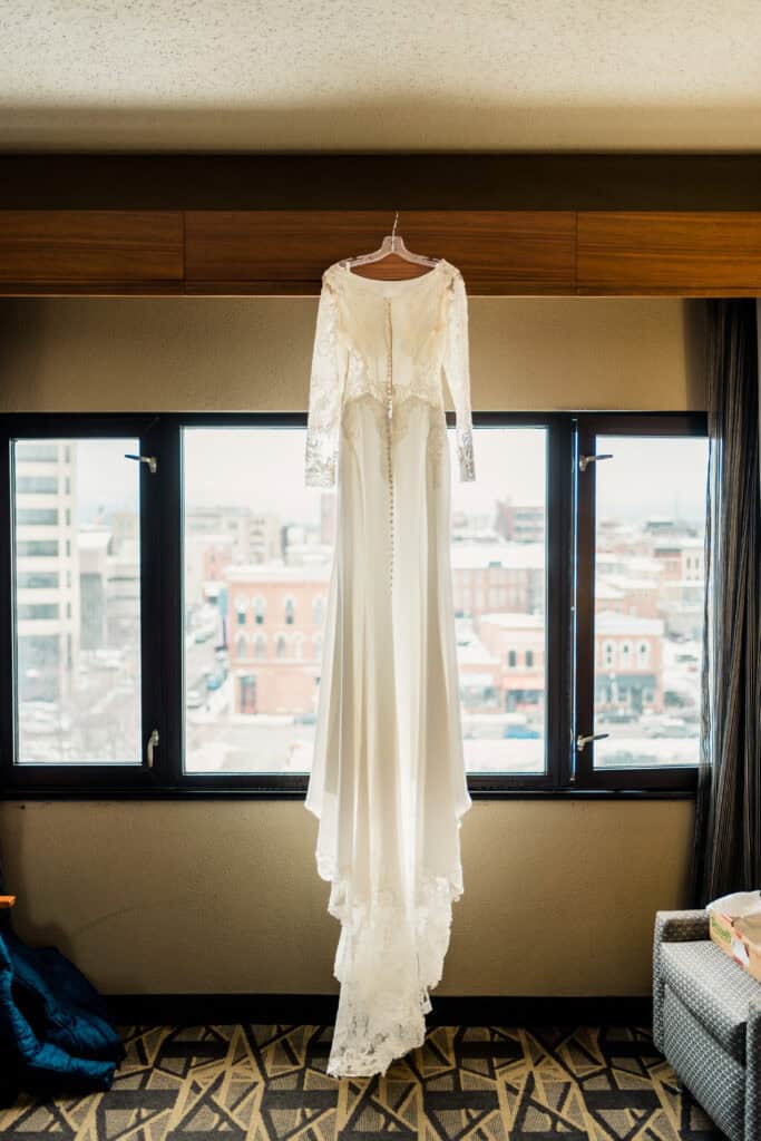 A long-sleeved white wedding dress with lace details is hanging on a wooden rod in front of a window, with a cityscape visible outside.